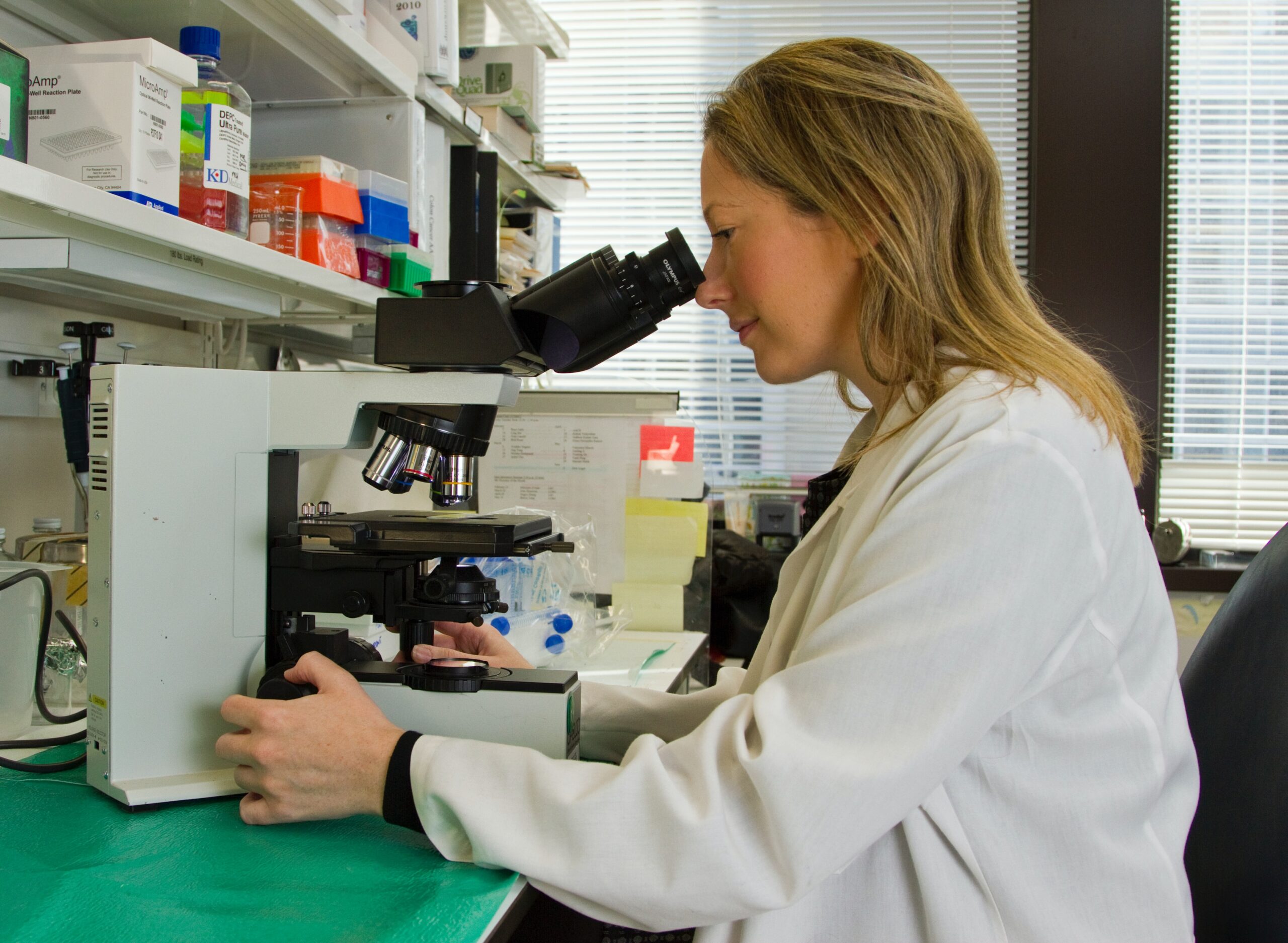 Blonde lady in a lab wearing a lab coat looking into a microscope